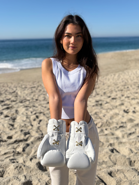 Woman posing on a beach wearing white shoes, pants, and gold accented, white boxing gloves. 